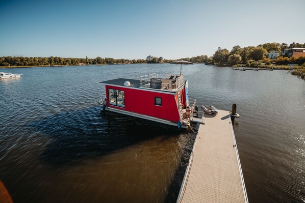 Una casa flotante roja amarrada a un muelle, con vistas claras al agua y una orilla boscosa al fondo.
