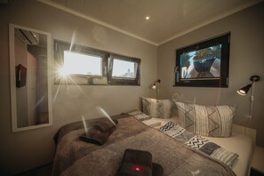 Un dormitorio con una cama grande, dos ventanas, un televisor montado en la pared y una lámpara de noche.
