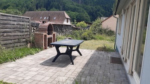 A patio with a table and a brick oven, surrounded by a wooden fence and a stone wall, with a forested hill in the background.
