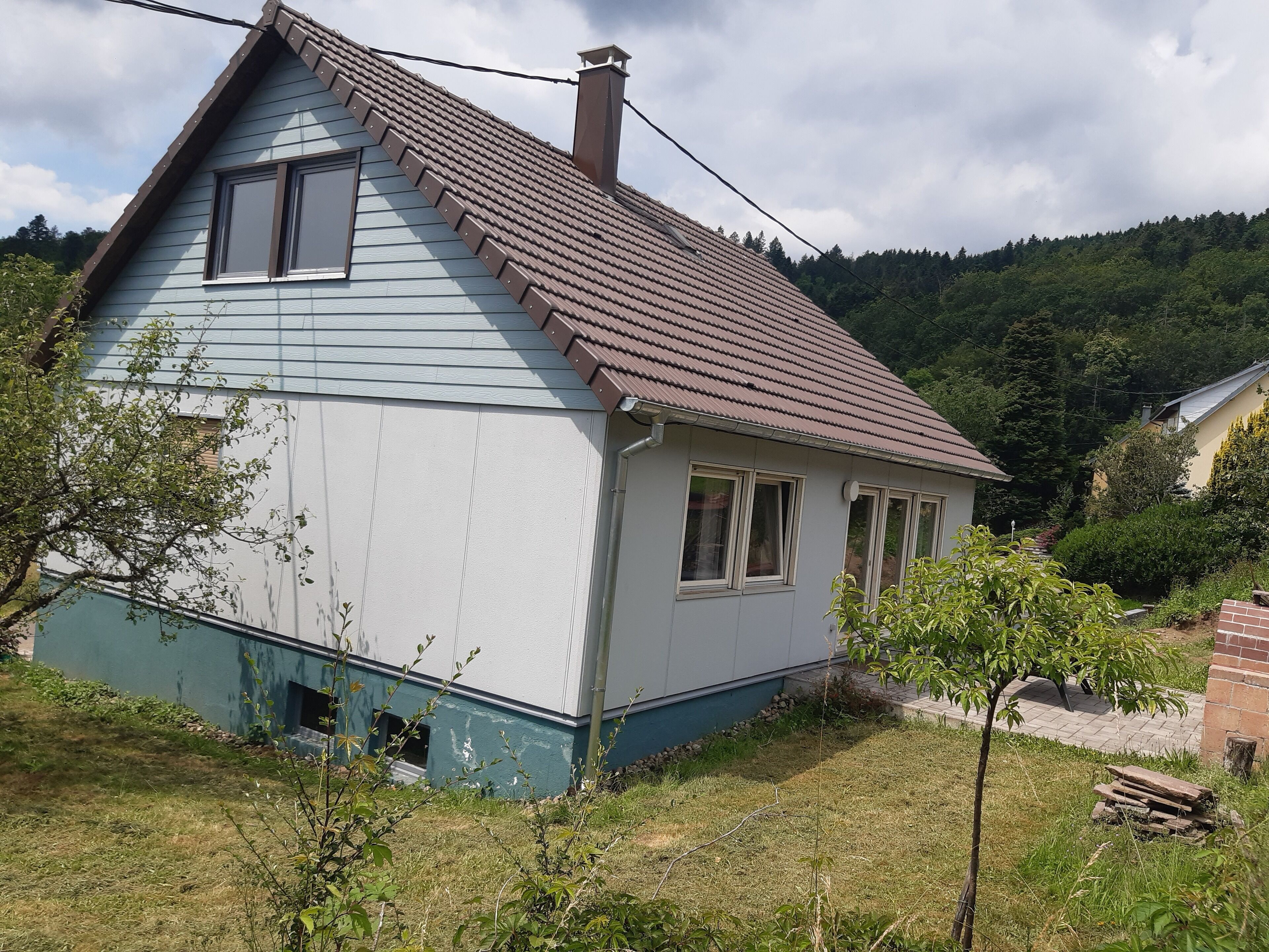 A small, single-story house with a sloped roof, surrounded by greenery and a hilly landscape.