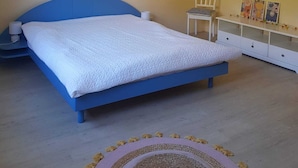 A bedroom with a blue bed, a white bedside table, a chair, a lamp, and a painting on the wall.