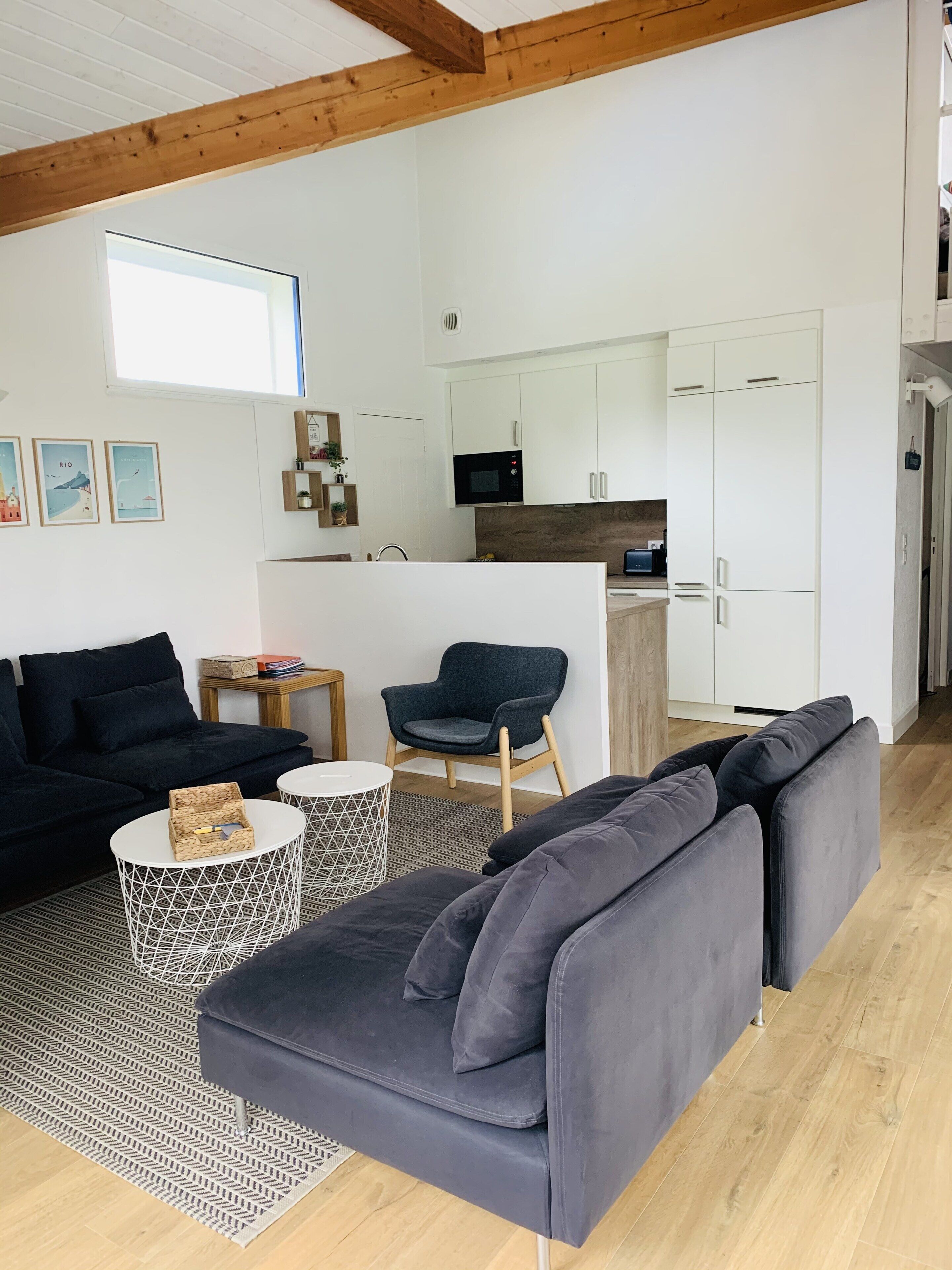 A modern living room with a dark grey sofa, a black armchair, and a white coffee table. There is a kitchen area with white cabinets and a microwave.