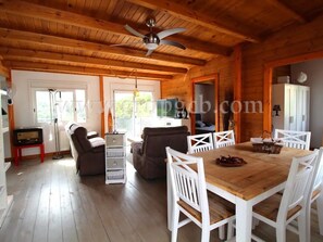 Interno di una baita in legno con un tavolo da pranzo, sedie bianche, una ventola a soffitto e un caminetto.