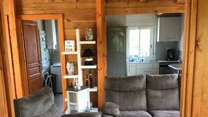 A cozy cabin interior with a wooden ceiling, a ceiling fan, a sofa, a dining table, and a kitchen area visible through an open door.