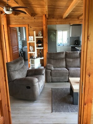 Interno accogliente di una baita con soffitto in legno, una ventola a soffitto, un divano, un tavolo da pranzo e una zona cucina visibile attraverso una porta aperta.