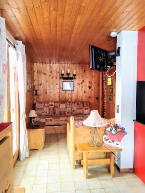 A cozy wooden cabin interior with a sofa, a wooden table with a lamp, and a television mounted on the wall.