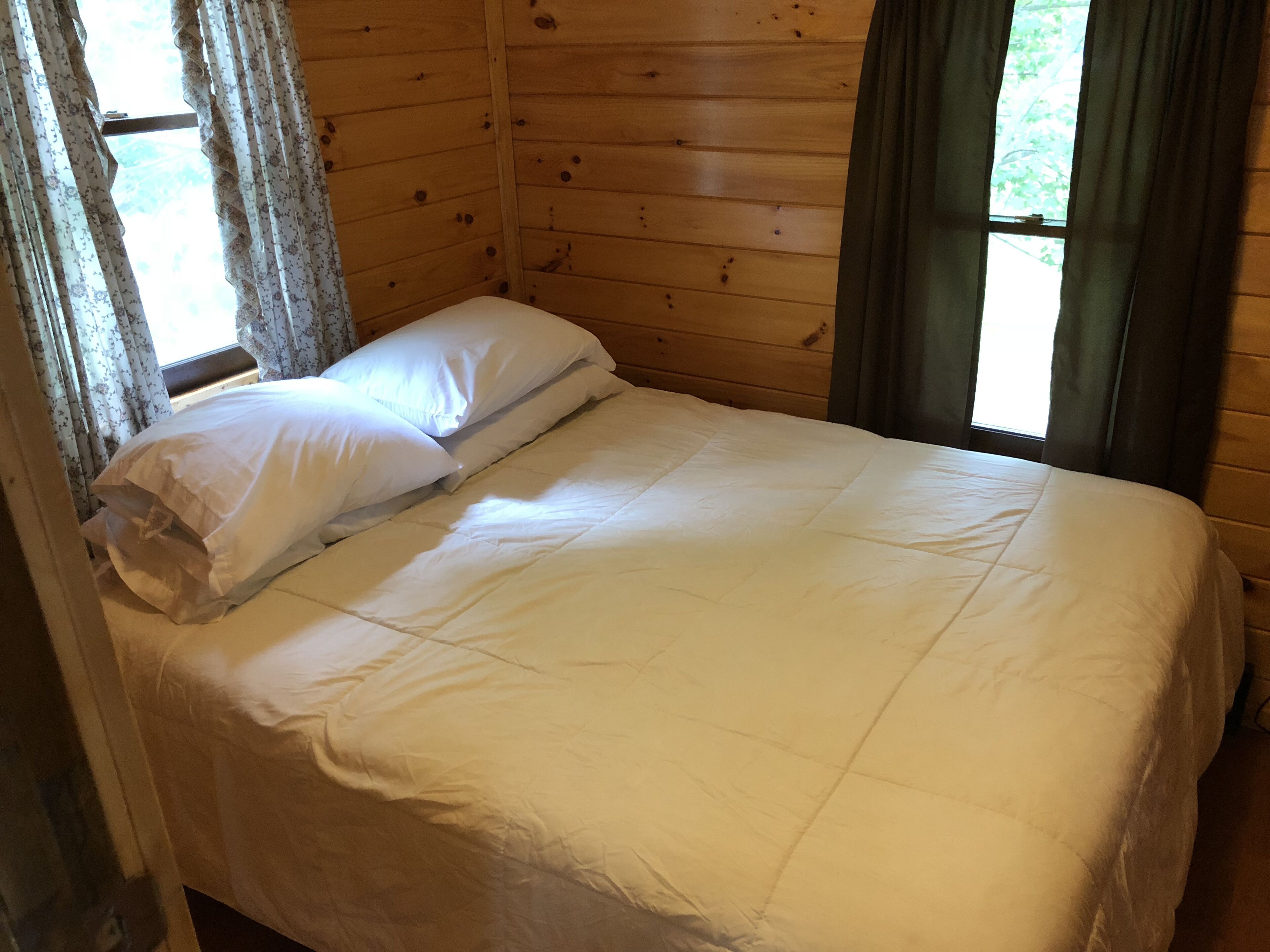 Un lit simple avec une literie blanche, dans une pièce aux murs en bois et dotée d’une fenêtre avec des rideaux.
