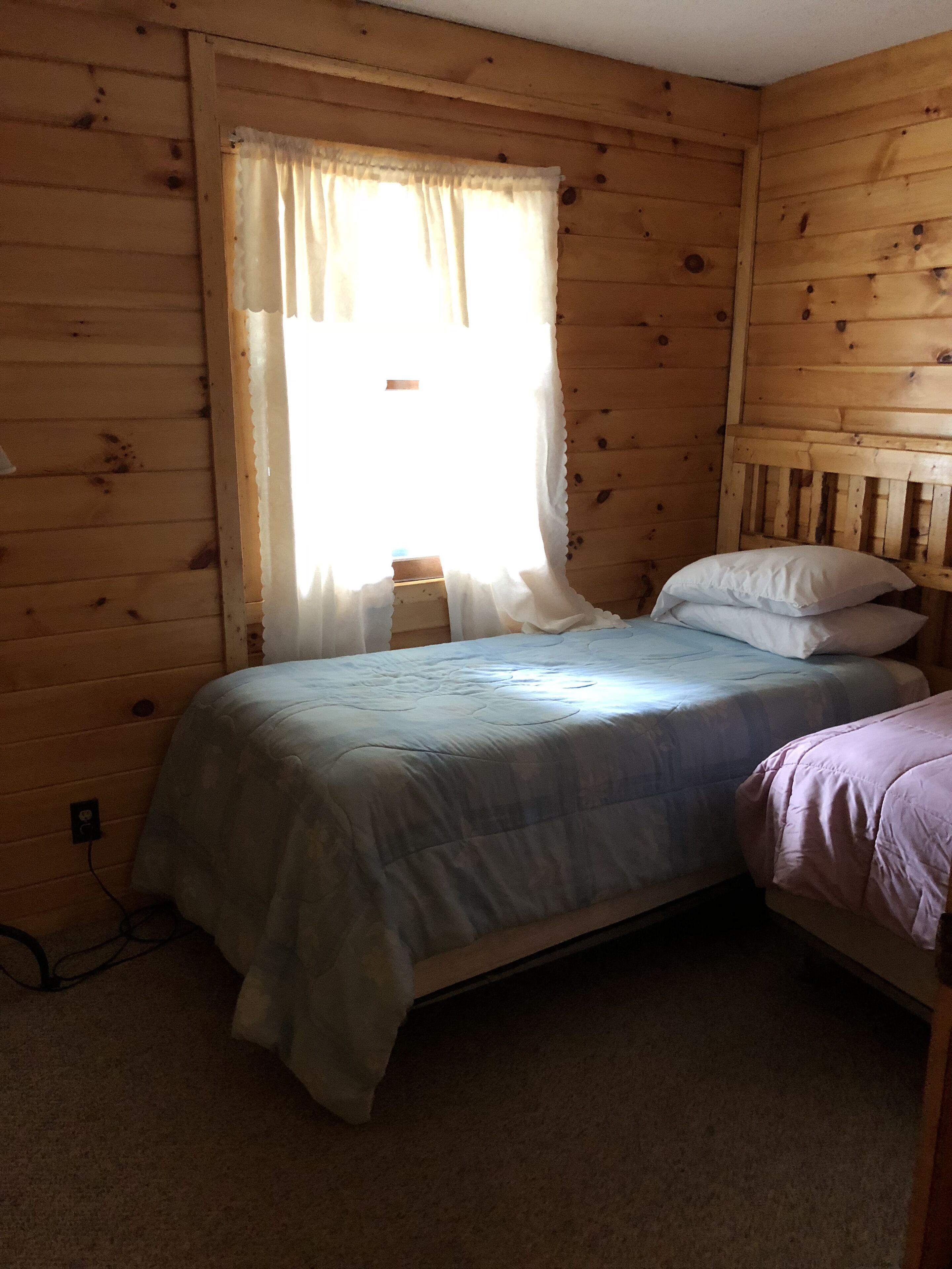 Pequeña habitación individual con cama, pared de madera y ventana con cortinas.
