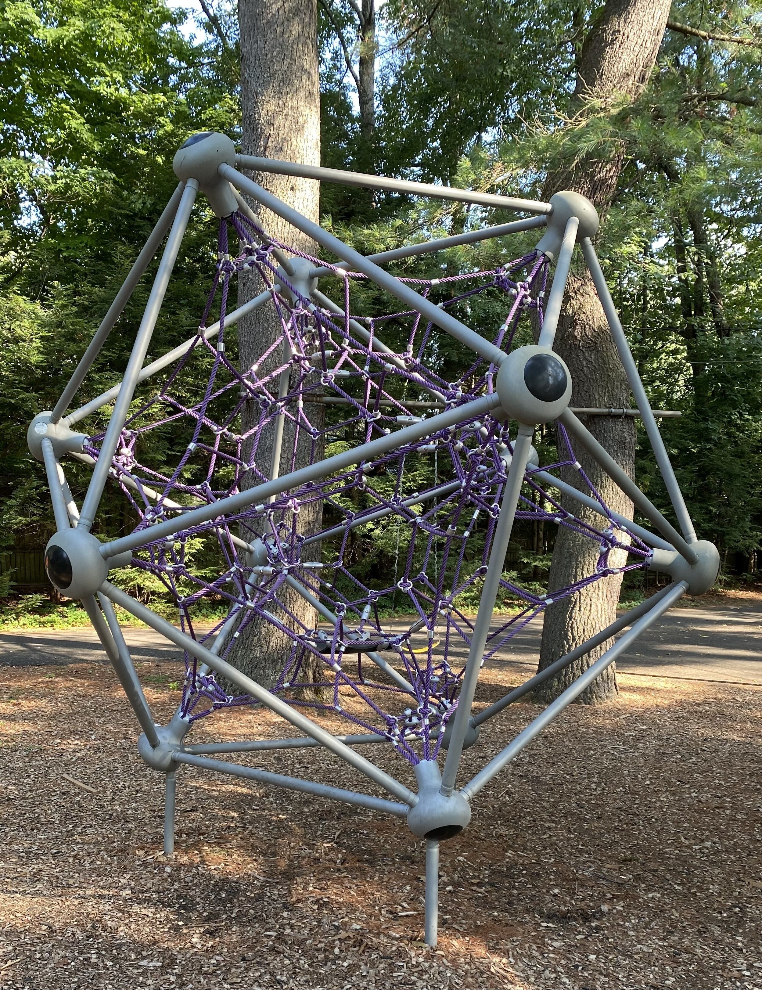 Eine geometrische Metallkonstruktion mit einem komplexen Seilnetz in einer bewaldeten Parkanlage.
