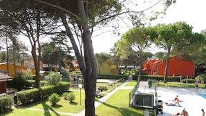 Uma área de piscina com zona para banhos de sol, espaço infantil e um caminho que leva a um edifício com fachada vermelha.