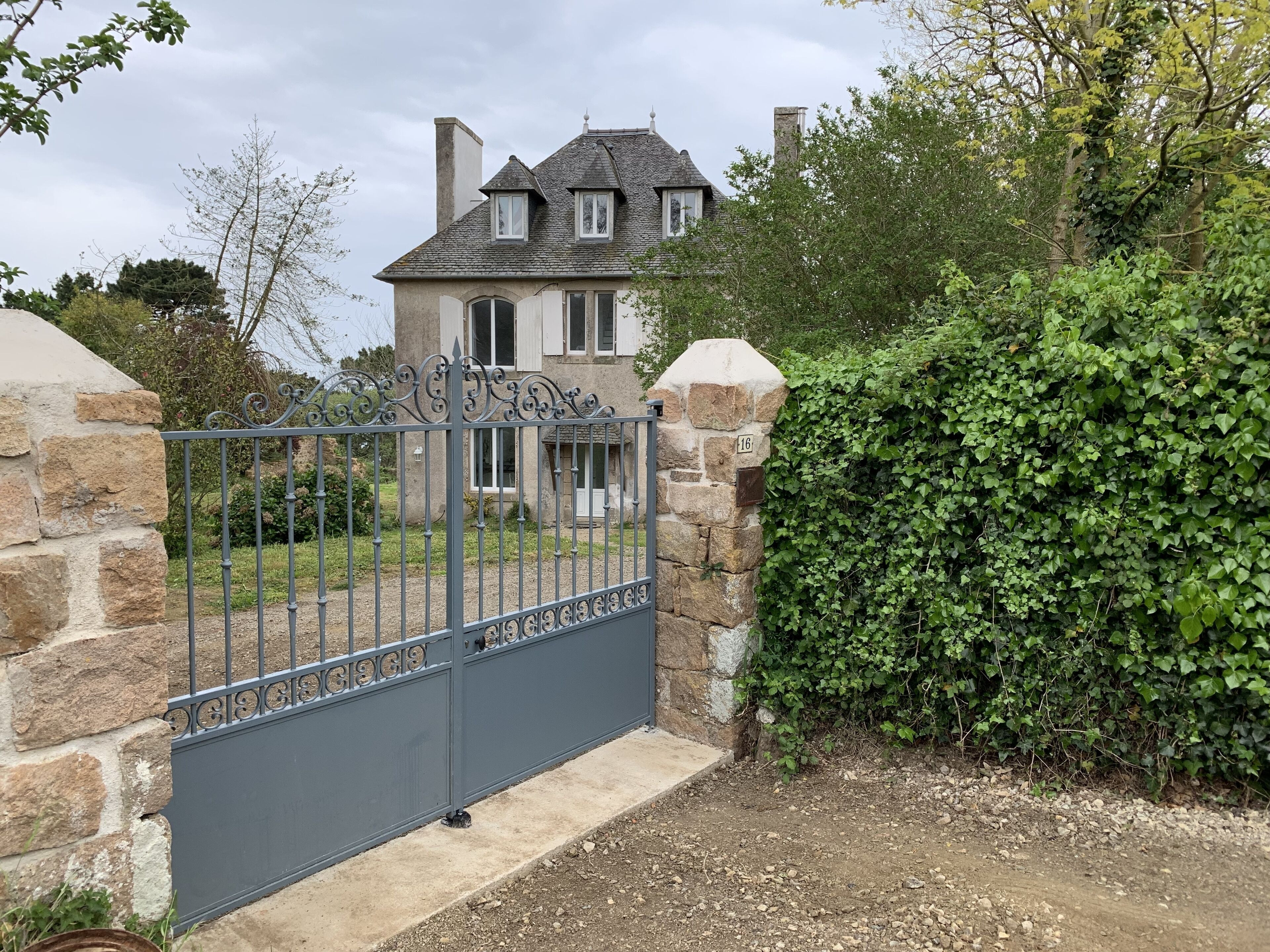 A stone house with a grey metal gate, surrounded by greenery and a stone wall.