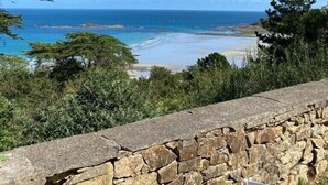 Vista costera con un muro de piedra y vegetación en primer plano, que conduce a una playa y al océano bajo un cielo despejado.