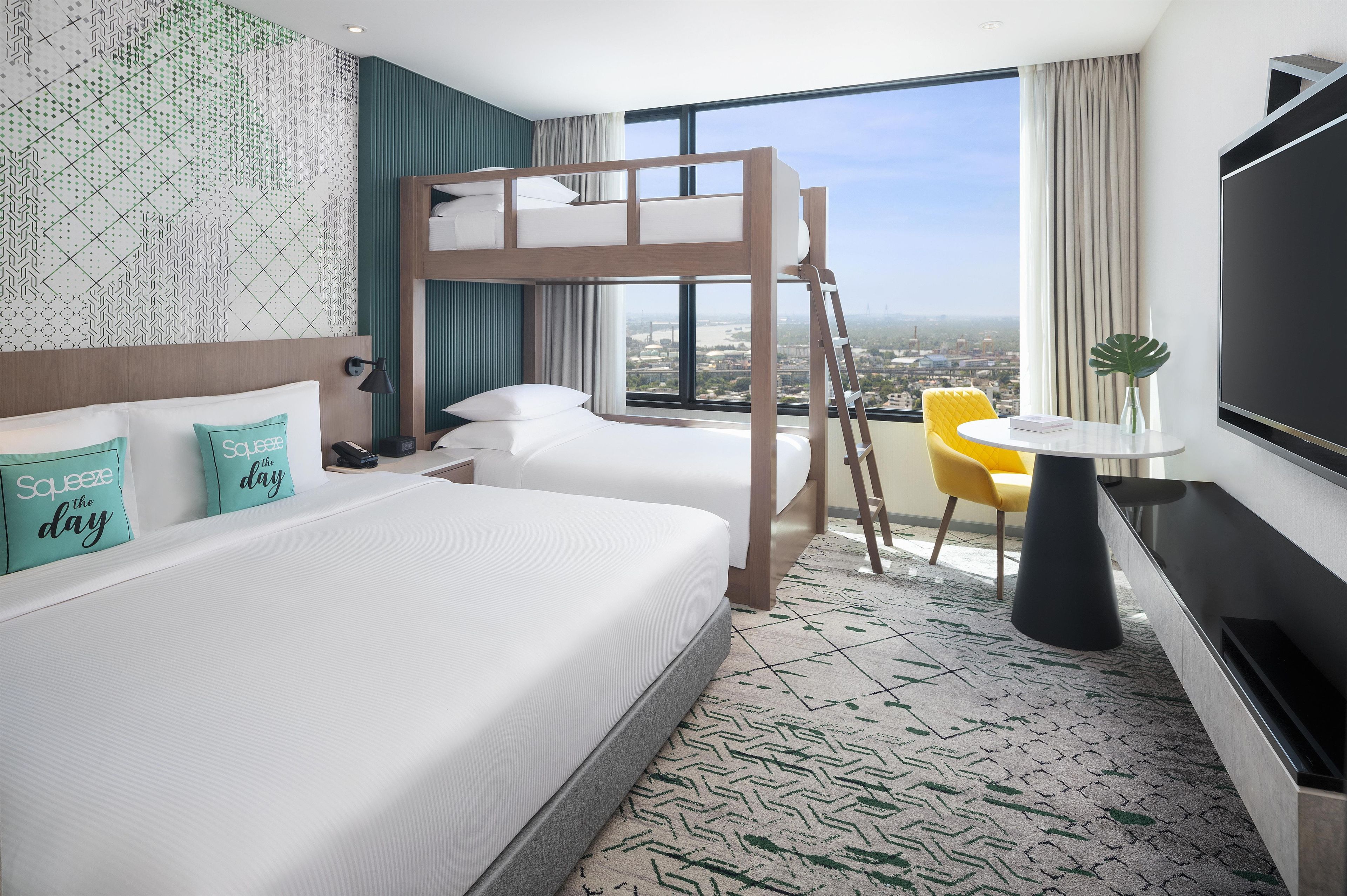 A hotel room with a bunk bed, a desk with a chair, a TV, and a view of the city.