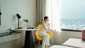 Ein Hotelzimmer mit Blick auf die Stadt, ein Schreibtisch mit Lampe, ein Sessel und ein kleiner Tisch mit Pflanze und Obst.