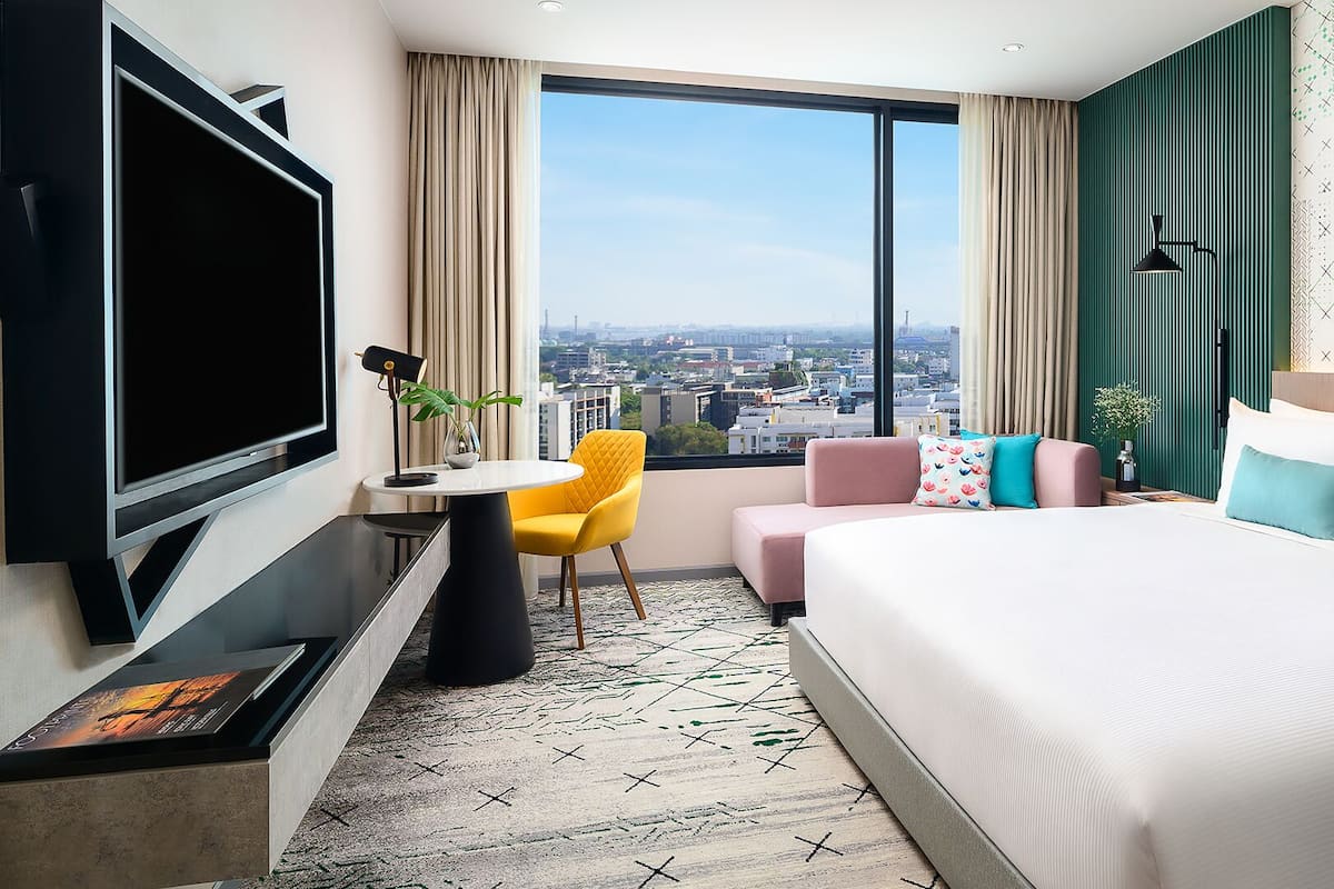 Une chambre d’hôtel moderne dotée d’un grand lit, d’un bureau avec une chaise, d’une télévision et offrant une vue sur la ville grâce à de grandes fenêtres.