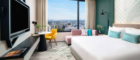 A modern hotel room with a large bed, a desk with a chair, a TV, and a view of the city through large windows.