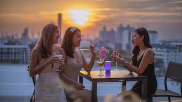 Trois femmes sont assises à une table, devant des boissons, et contemplent un paysage urbain au coucher du soleil.