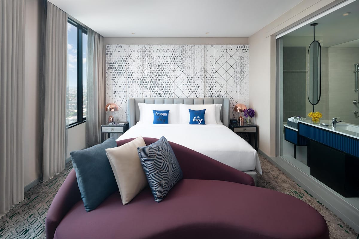 Une chambre d’hôtel moderne dotée d’un grand lit, d’un canapé séparé, d’une salle de bain avec un grand miroir et offrant une vue sur la ville.