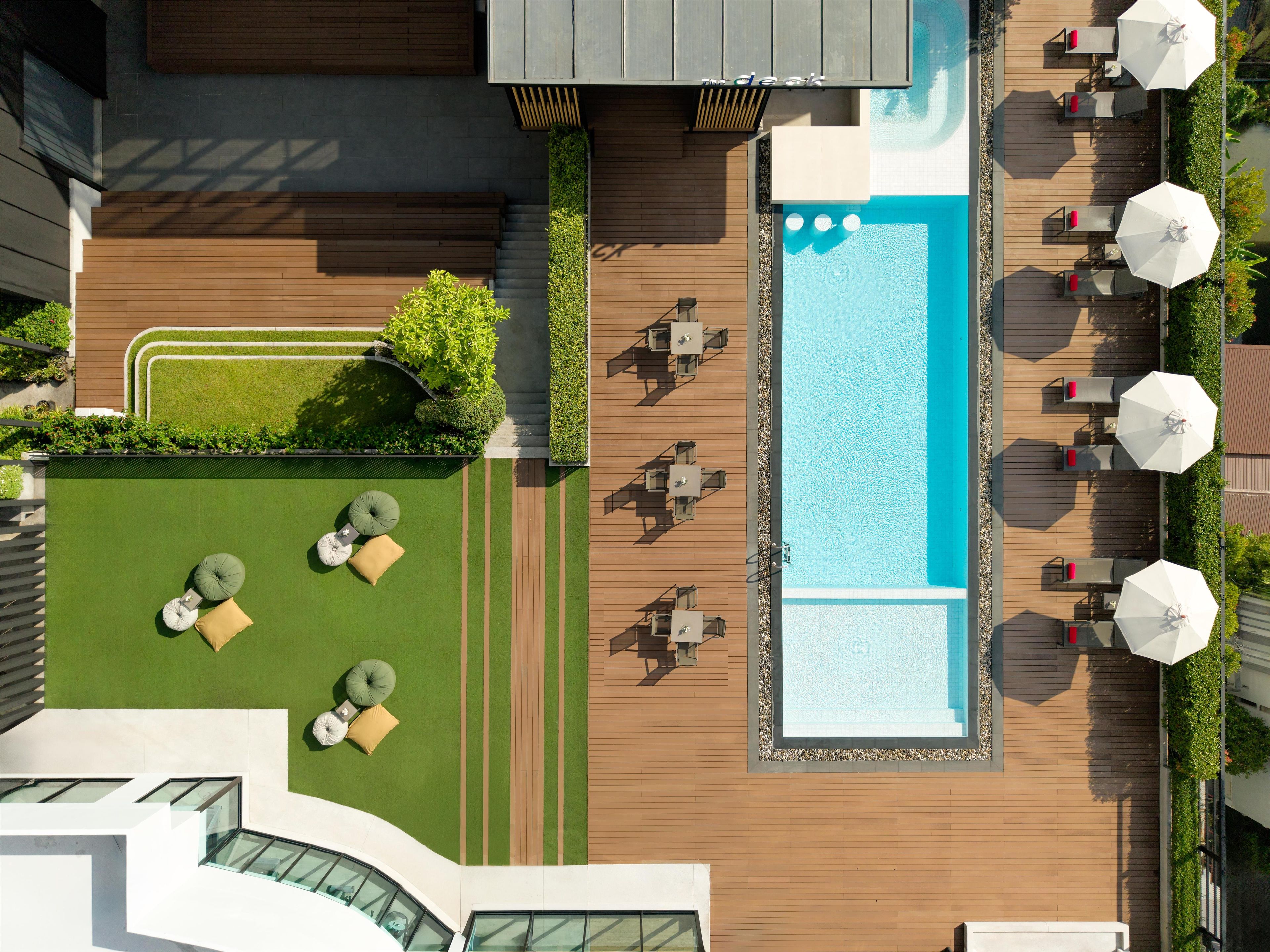 Un área de piscina en la azotea con tumbonas y sombrillas, rodeada por una terraza de madera y un pequeño jardín.