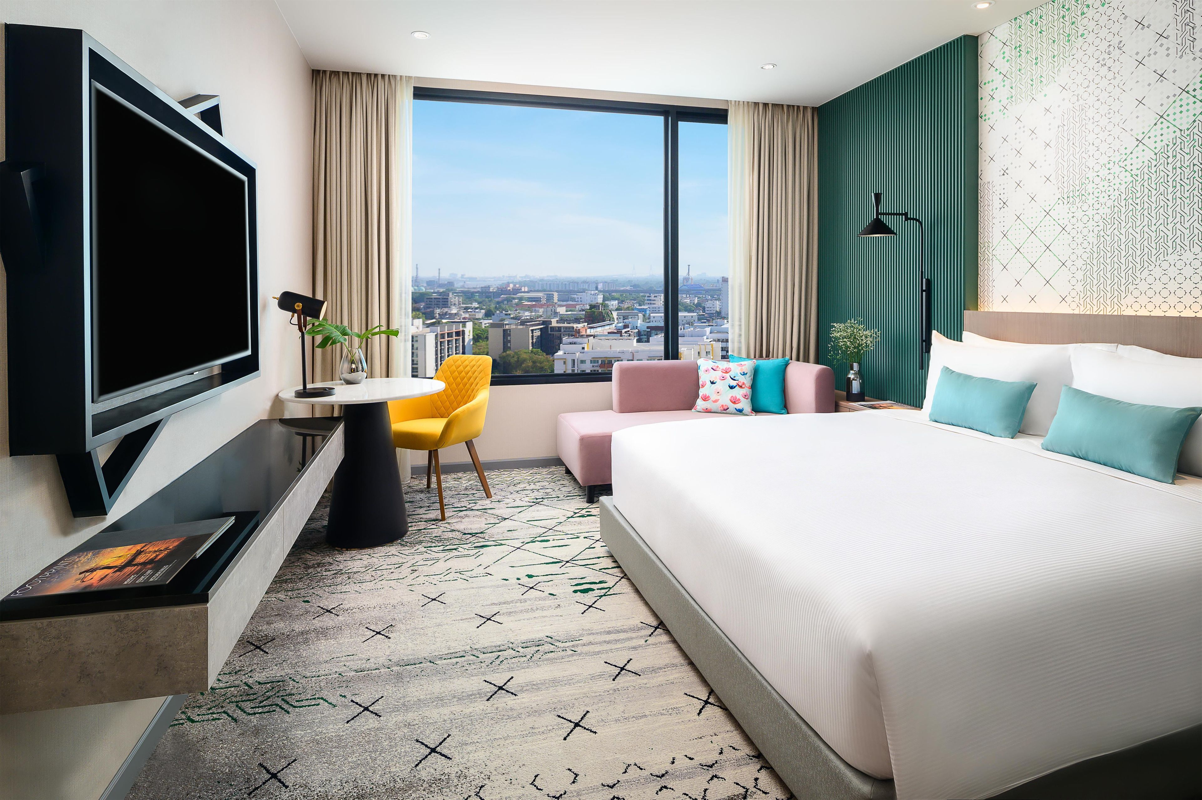 A hotel room with a large bed, a TV, a desk with a chair, and a view of the city.
