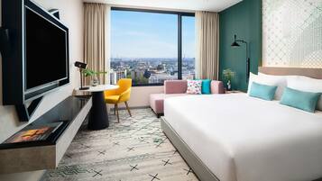 A hotel room with a large bed, a TV, a desk with a chair, and a view of the city.