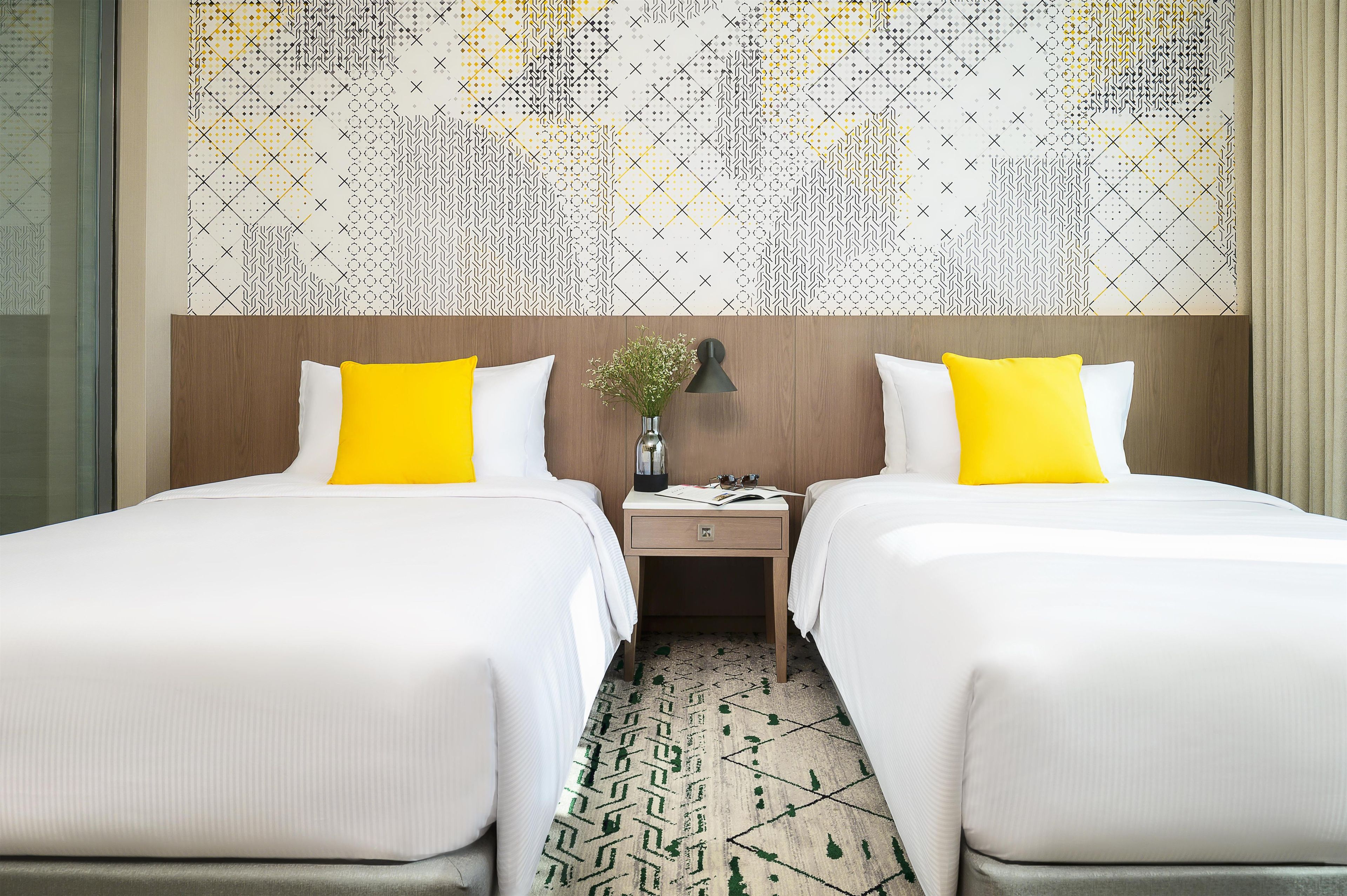 A hotel room with two beds, a nightstand, a vase with flowers, and a patterned wall.
