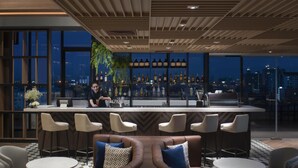 Een moderne bar met uitzicht op de stad, barstoelen en een zithoek met een geruit vloertapijt.