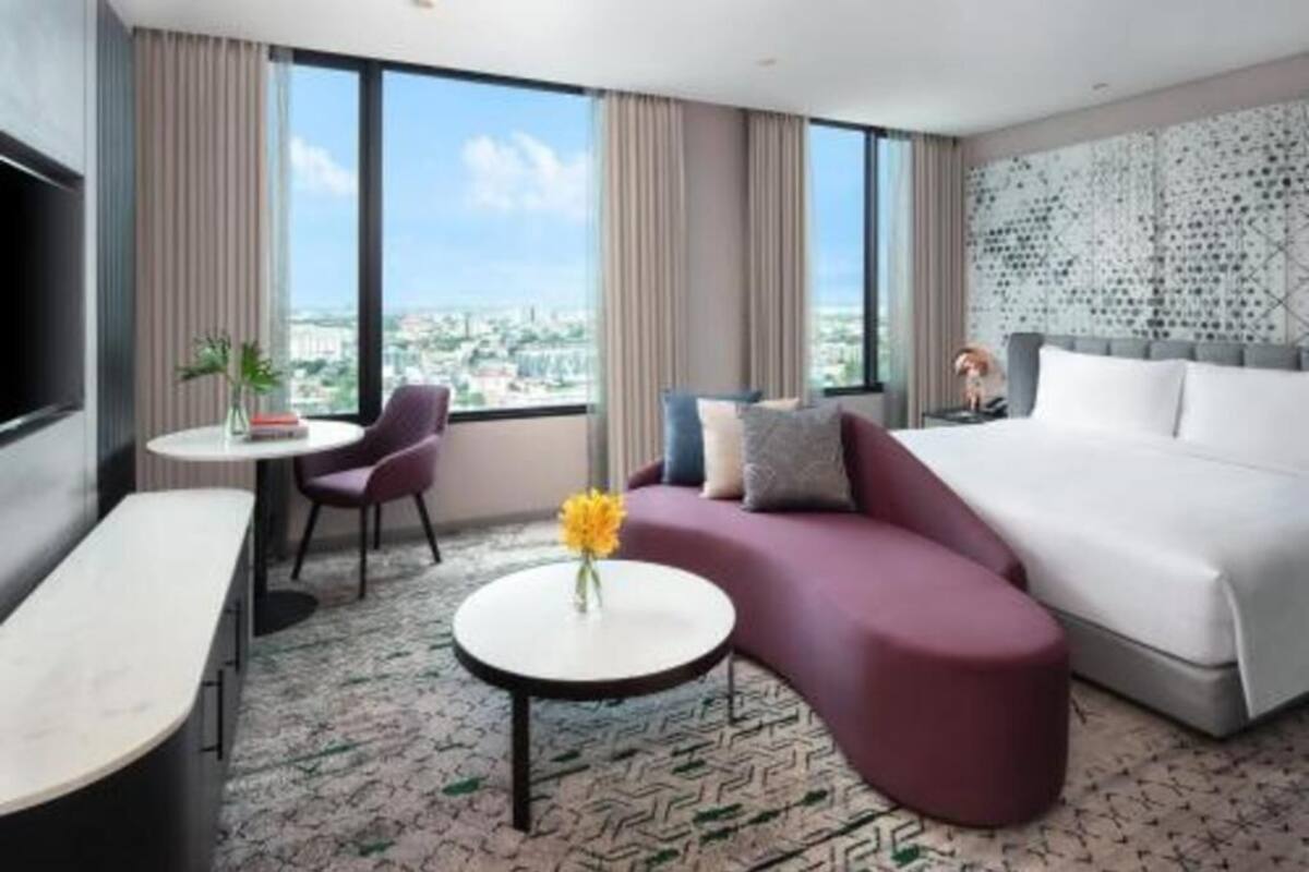 Une chambre d’hôtel moderne avec un canapé incurvé, un lit, une petite table ronde ornée d’un vase et de grandes fenêtres donnant sur la ville.