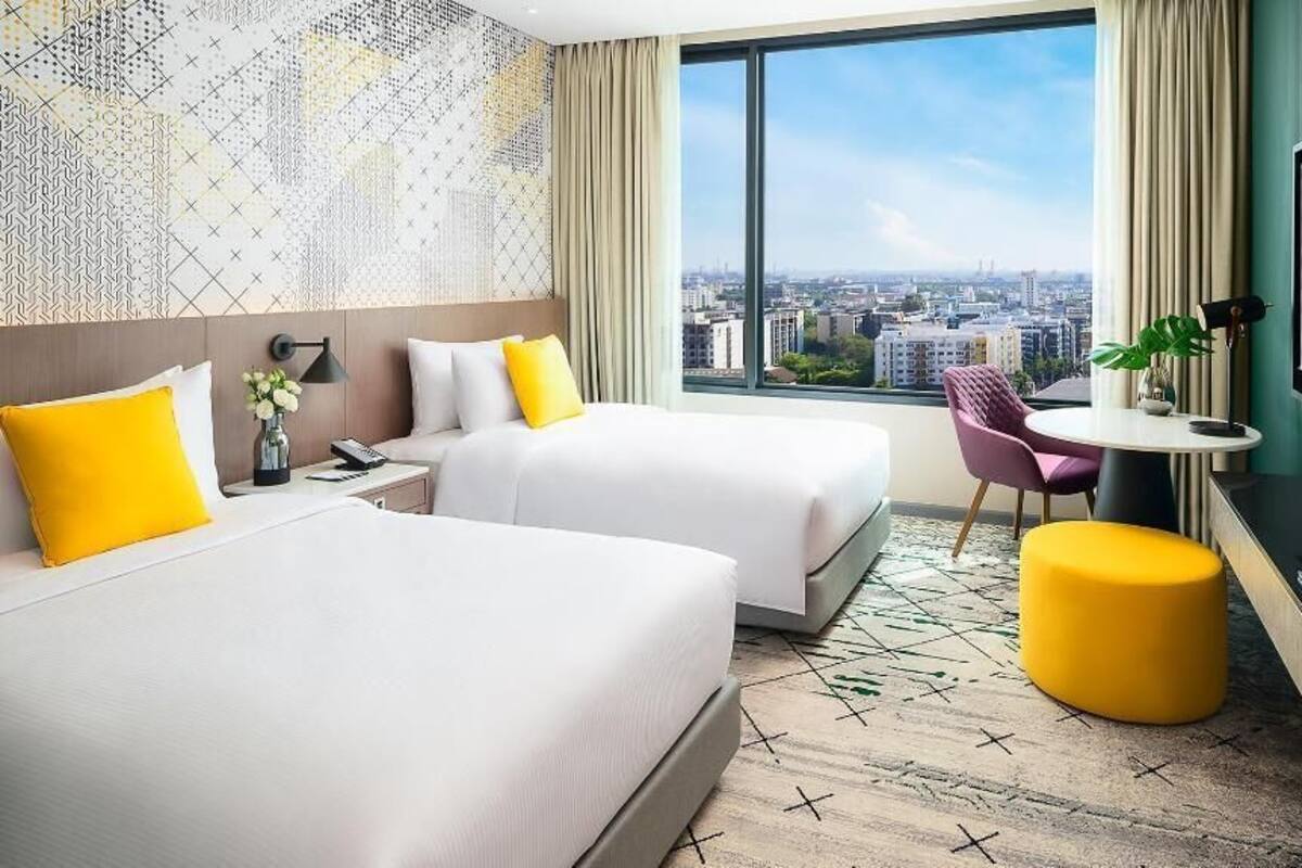 Une chambre d’hôtel avec deux lits, une télévision, une petite table, une chaise et une grande fenêtre donnant sur la ville.