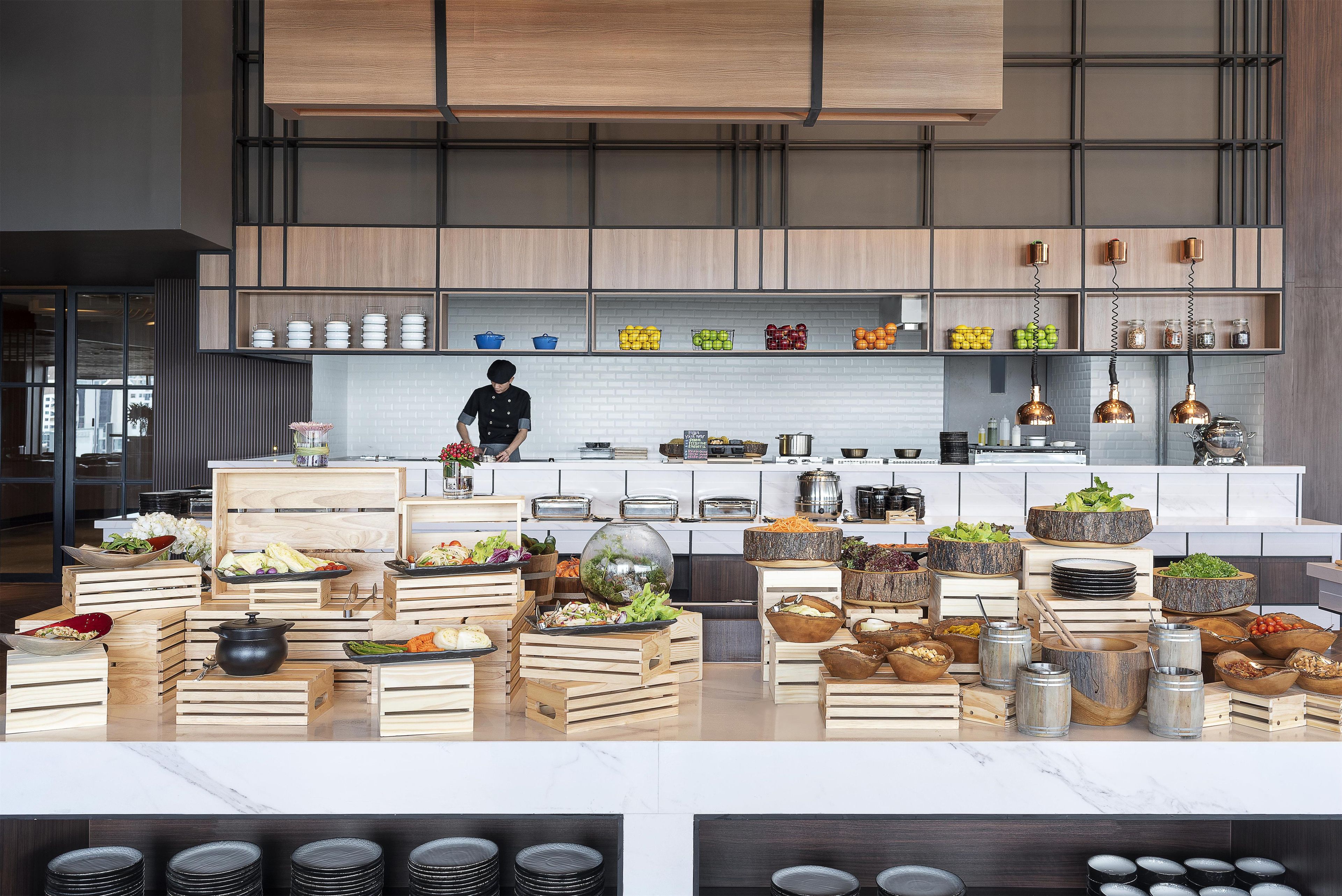A modern kitchen with wooden cabinetry, a chef preparing food, and various food items displayed on wooden crates.
