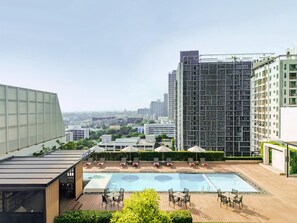 Un espace piscine aménagé sur le toit, avec des chaises longues et une piscine à ciel ouvert protégée par des parois de verre, le tout entouré de bâtiments modernes et de verdure.