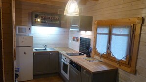 A compact kitchen with wooden walls, a microwave, a refrigerator, a sink, and a window with decorative curtains.