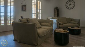 A spacious living room with a large window, a clock on the wall, and a ceiling light fixture. - Mas Provençal first floor living room with fireplace and veranda overlooking Alpilles- Vallée des Baux (Paradou)
