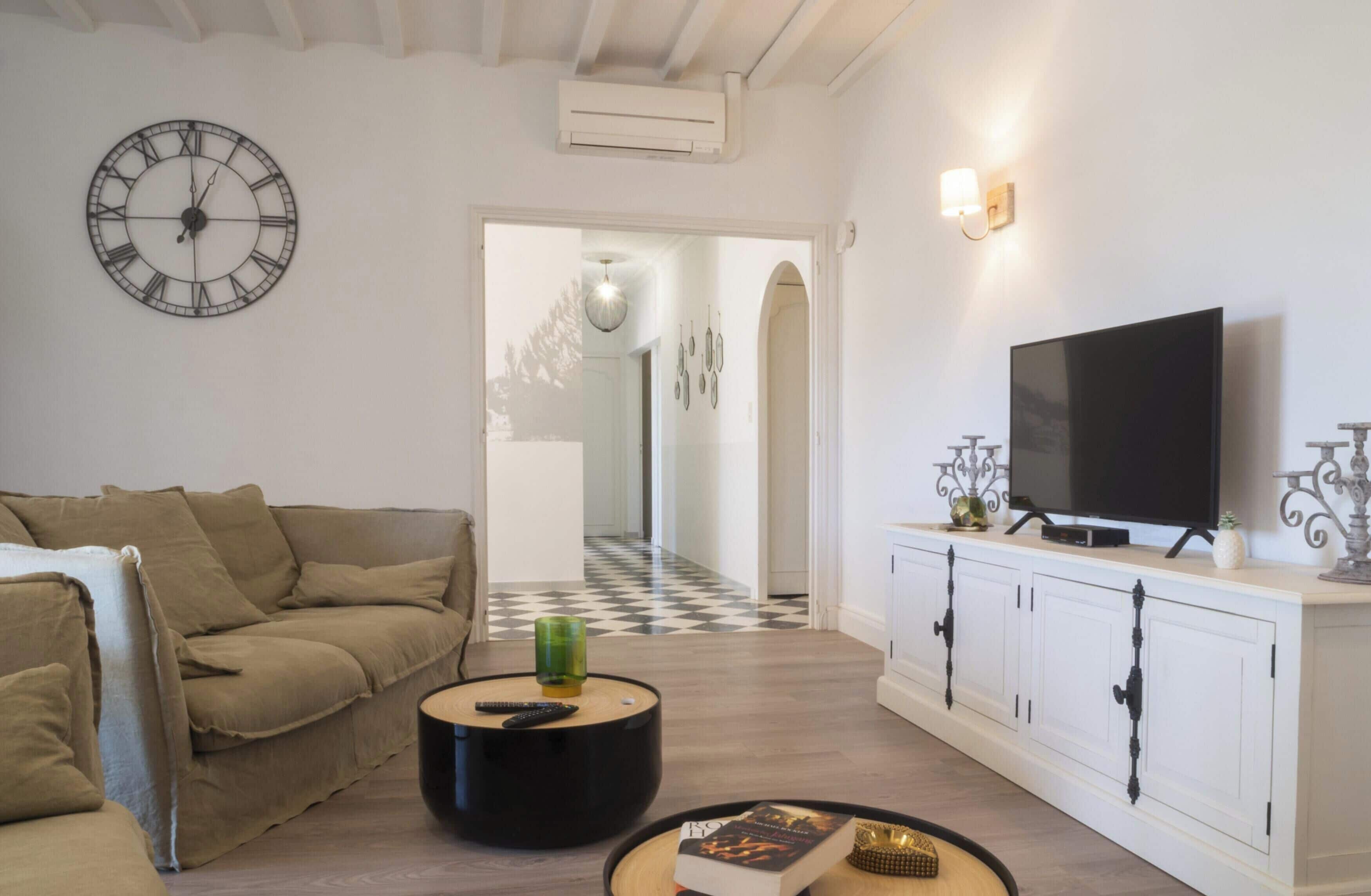 Uma sala de estar com sofá, mesa de centro com um livro, televisão em um móvel e relógio de parede.