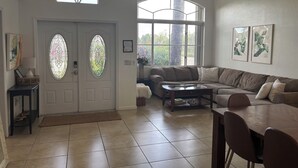 Ein geräumiges Wohnzimmer mit einer großen Eckcouch, einem Couchtisch und einem Esstisch mit Stühlen. Zwei große Fenster bieten natürliches Licht und Blick auf Grünfläche.