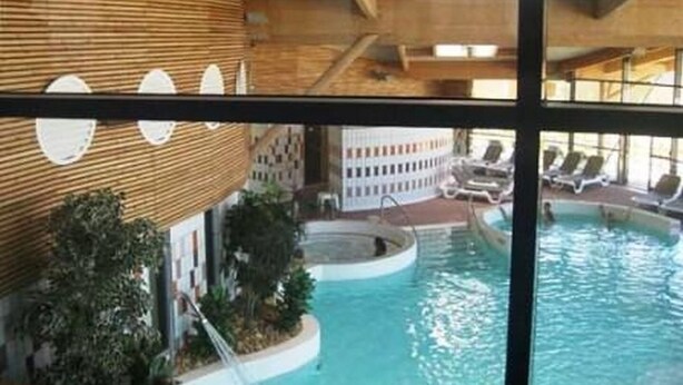 Une piscine intérieure avec un plafond en bois, d’où l’on peut apercevoir des chaises longues et un bâtiment au loin.