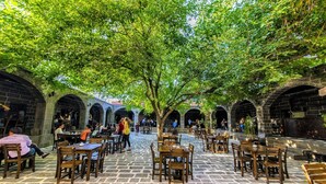 Un café en plein air doté d’un sol en pierre et de tables et chaises en bois, entouré d’arcades en pierre et d’arbres.