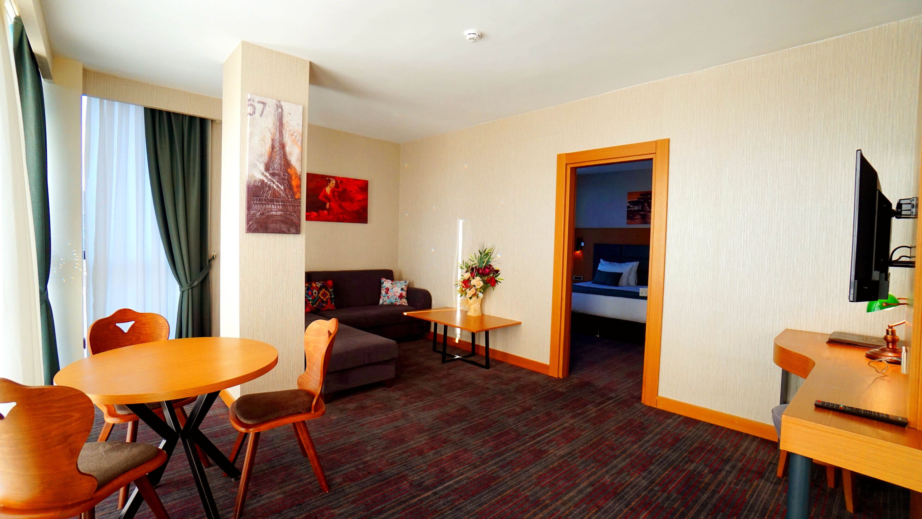 A hotel room with a dining table, chairs, a sofa, a small table with a vase of flowers, a bed, and a television mounted on the wall.