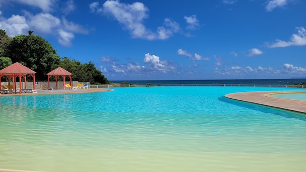 A clear blue pool with lounge chairs and red gazebos, surrounded by lush greenery and a view of the ocean. - Anse des rochers - Studio Cashew (saint-françois)