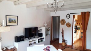 A living room with a TV, speakers, and a chandelier. There is a dining table with a vase of flowers. A kitchen is visible through an open door.