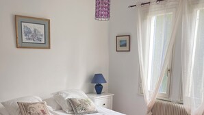 A neatly made bed with a brown bedspread, two rolled towels, white pillows, and a blue bedside lamp on a white nightstand.