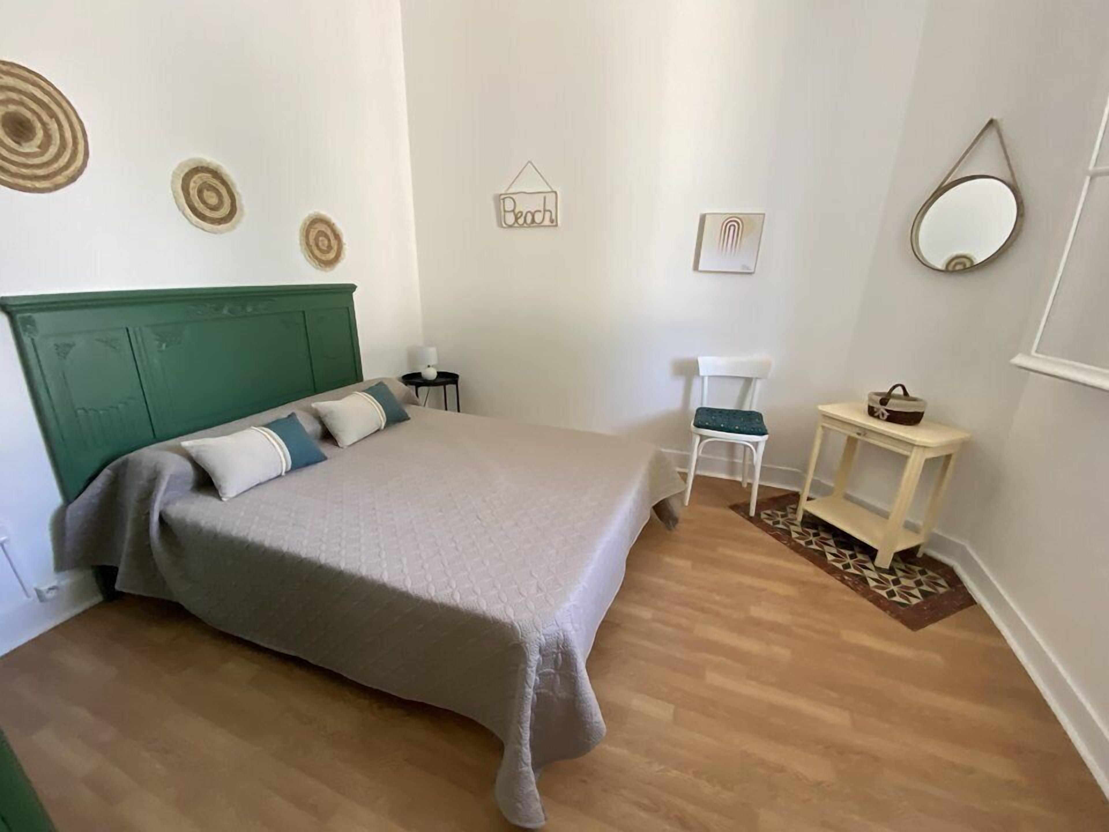 Ein Schlafzimmer mit einem grünem Kopfteil, einem Bett mit grauer Decke, einem weißen Stuhl, einem kleinen Tisch mit Spiegel und Wanddekorationen, darunter ein Schild mit der Aufschrift 'Beach' und runde Muster.