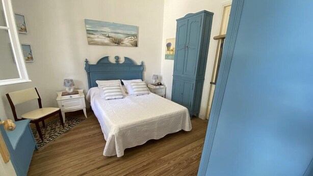 Un dormitorio con piso de madera, una cama con ropa de cama blanca, una mesita de noche, una silla y un armario.