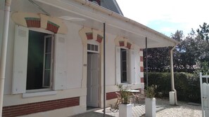 A building with a white facade, red brick base, and multiple windows with shutters. There is a covered outdoor area with a table and chairs.