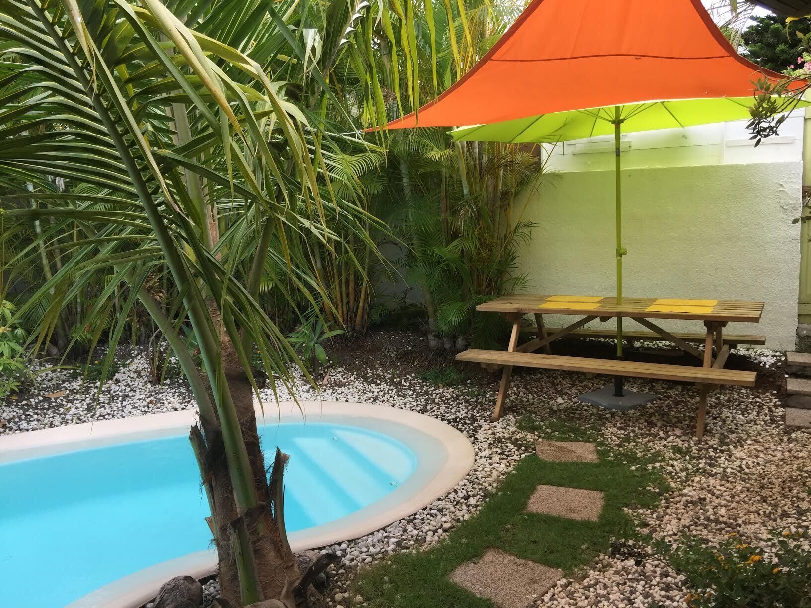 Área à beira da piscina com um banco de madeira, um guarda-sol laranja e uma pequena piscina rodeada por pedras e vegetação.