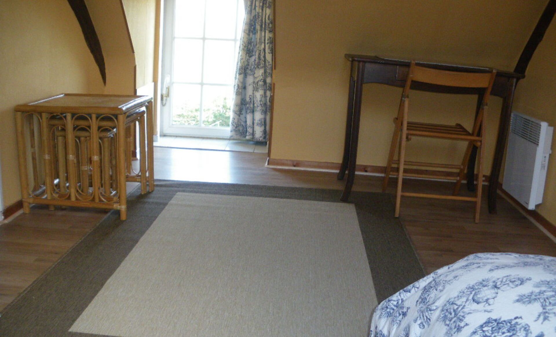 A room with a wooden bed, a small wooden table, a chair, and a large window with curtains.