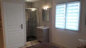 Un dormitorio con cama, mesita de noche, ventana con persianas, zona de ducha con mampara de cristal y baño con lavabo y espejo.