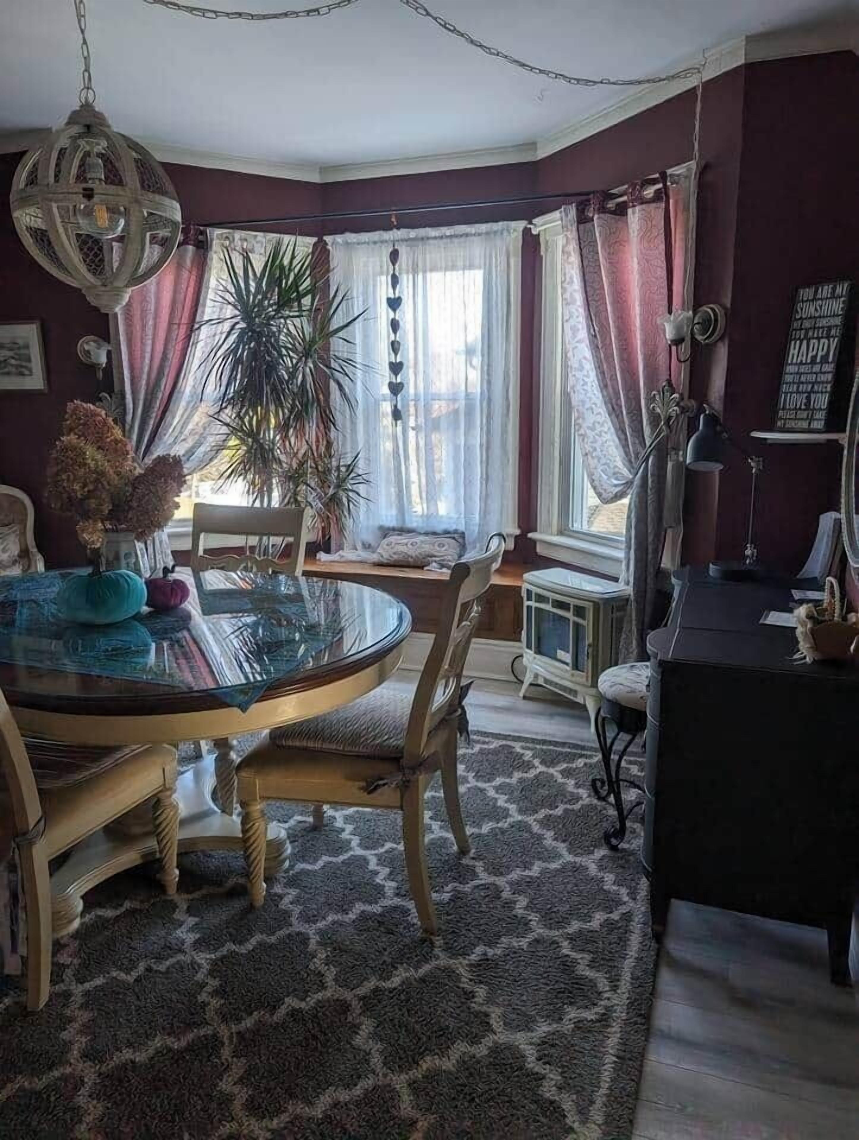 En matplats med ett runt glasbord, trästolar, en kristallkrona och ett stort fönster med gardiner.