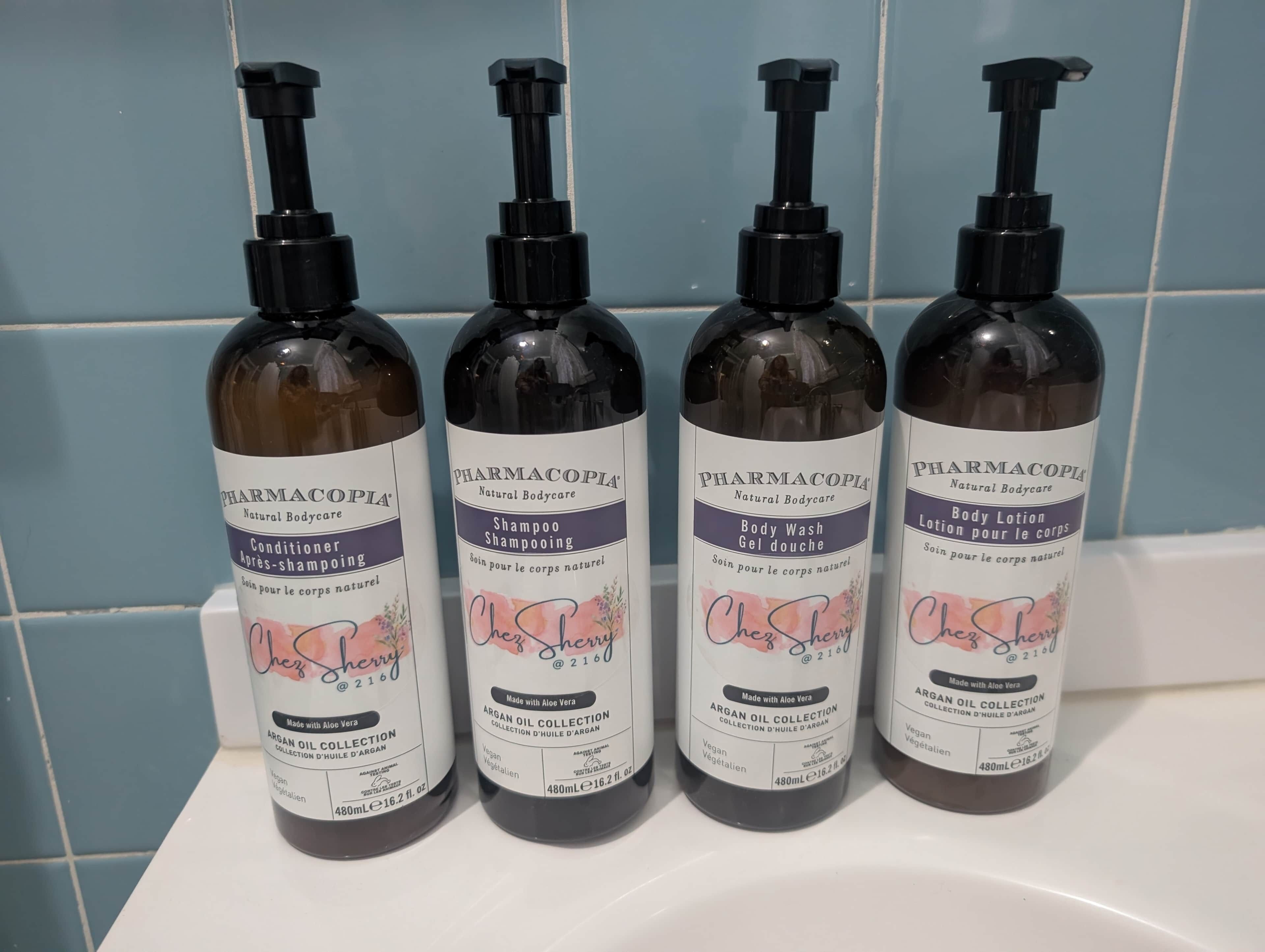Four bottles of body care products with pump dispensers, labeled 'PHARMACOPIA Natural Bodycare,' on a white surface against a blue tiled wall.