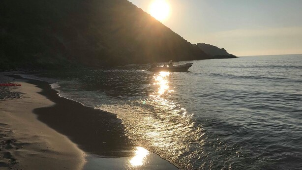 En strand med en båd, roligt vand og en solbeskinnet bakke i baggrunden.